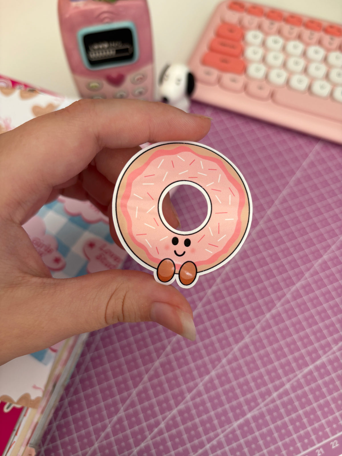 A hand holds a die-cut sticker of a cute pink donut with sprinkles and a smiling face. The background isn't fully in focus but shows a pink desk mat, a pink retro phone, a pink keyboard, and a notebook with pastel cloud stickers.