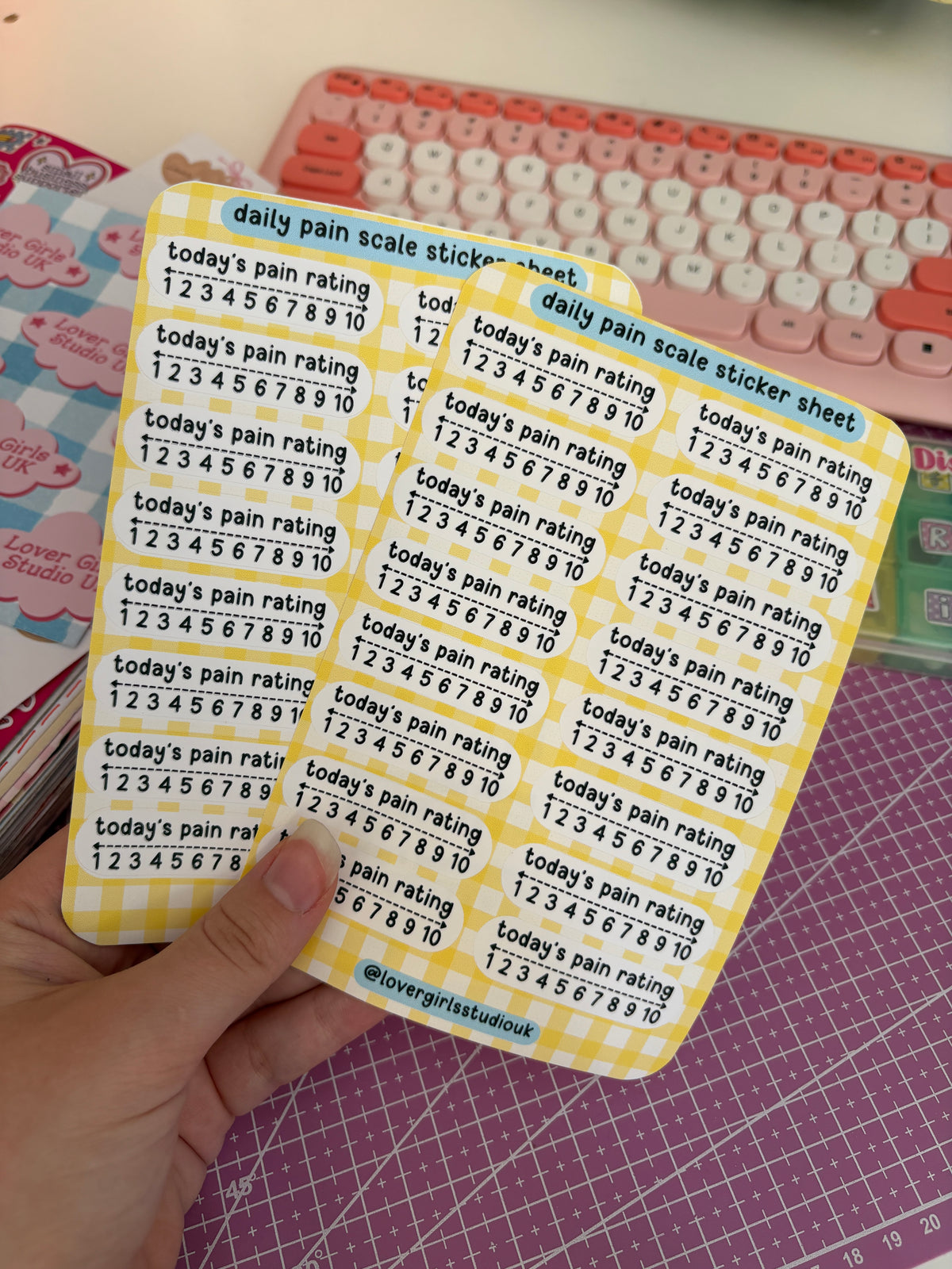 A hand holds a yellow gingham sticker sheet with rows of daily pain scale stickers numbered 1 to 10, with text reading 'today's pain rating 1 2 3 4 5 6 7 8 9 10'. A pink and white keyboard and a purple cutting mat are visible in the background.