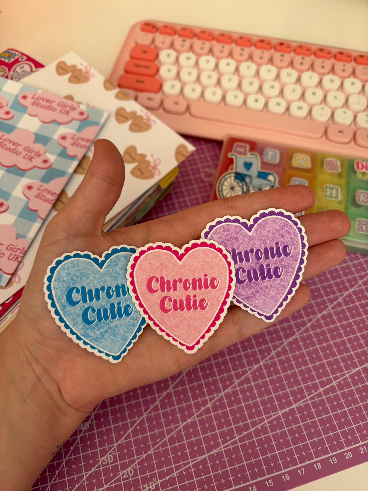 Three heart-shaped stickers with scalloped edges reading 'Chronic Cutie' in blue, pink, and purple, held in a person's hand over a pink cutting mat.