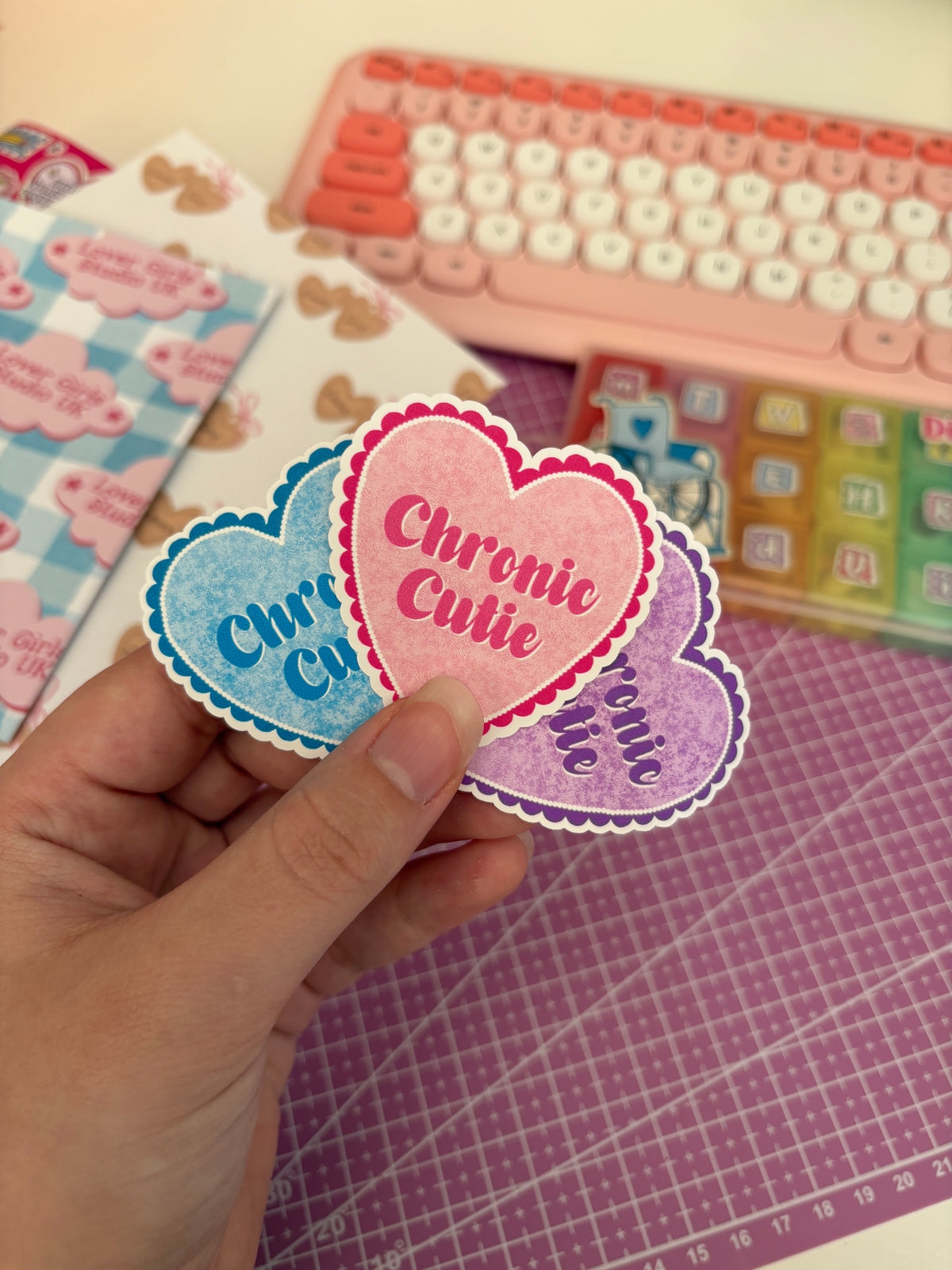A hand holds three heart-shaped stickers with scalloped edges reading 'Chronic Cutie' in pink, blue, and purple, with a pink keyboard and crafting tools blurred in the background.