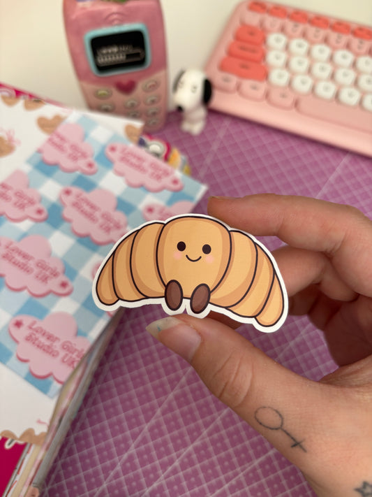 A hand holds a die-cut sticker shaped like a smiling croissant, with a pink desk background and pastel stationery items blurred behind it.