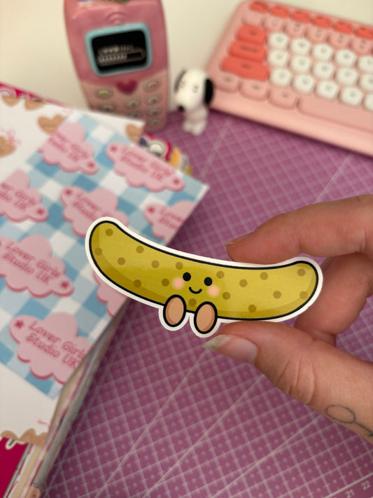 A hand holds a die-cut sticker of a cute green pickle and a smiling face. The background isn't fully in focus but shows a pink desk mat, a pink retro phone, a pink keyboard, and a notebook with pastel cloud stickers. 