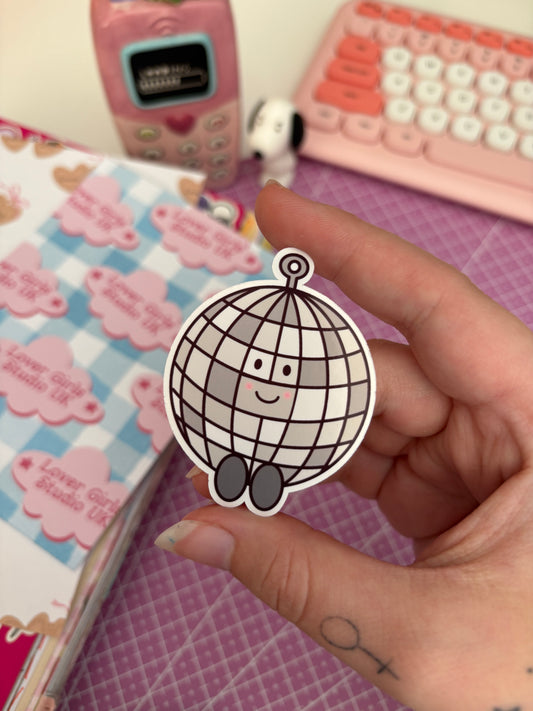 A hand holding a cute die-cut sticker of a smiling disco ball character, with a pink-themed notebook and keyboard in the background.